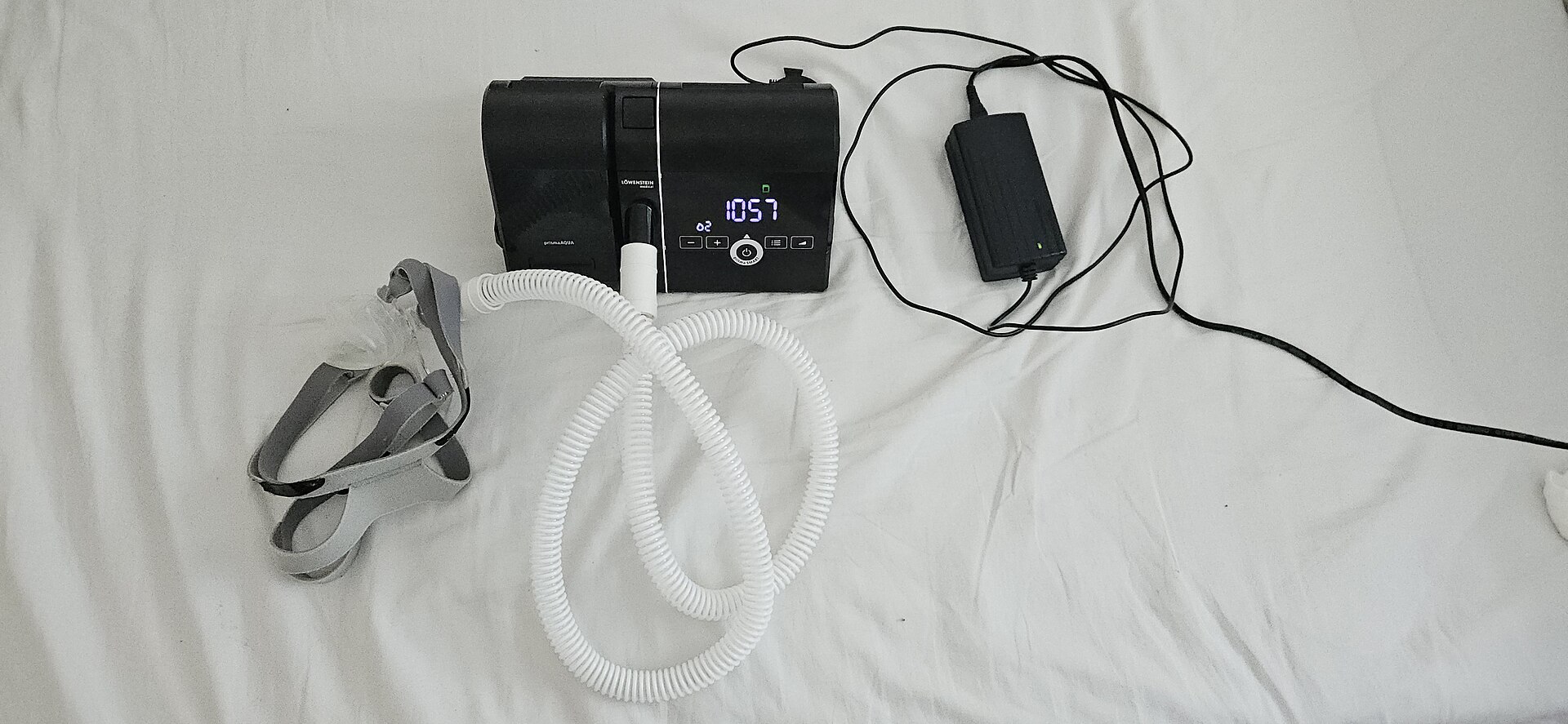A CPAP machine with its hose, mask and headgear on white bedsheets.