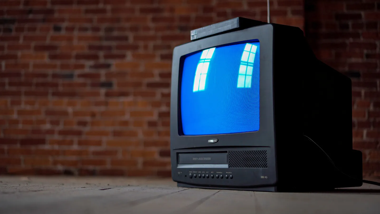 An old CRT television with a built-in VCR, its screen glowing blue, against a brick wall.