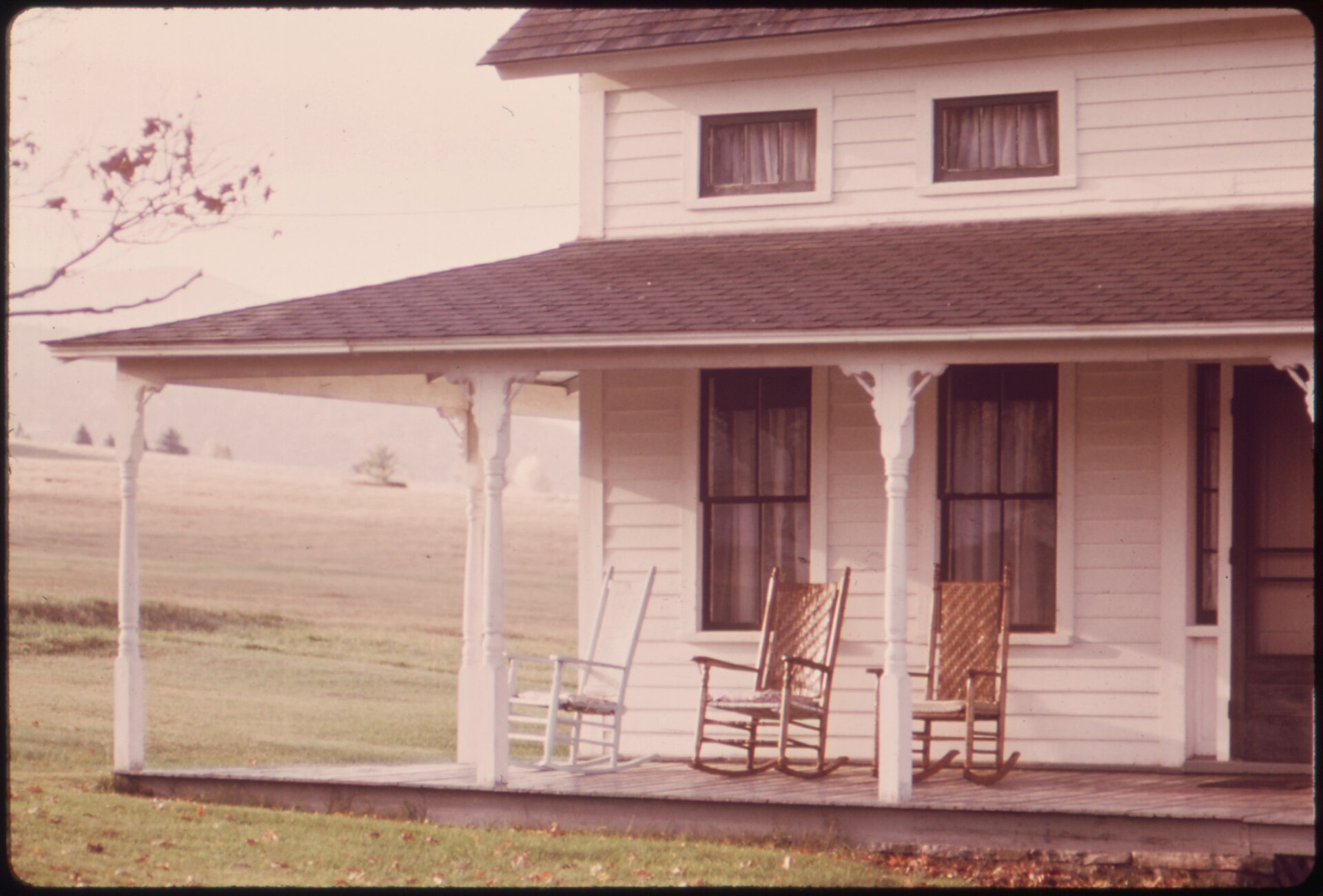 Three empty rocking chairs on the porch of a white clapboard farmhouse, in a faded 1970s photograph.